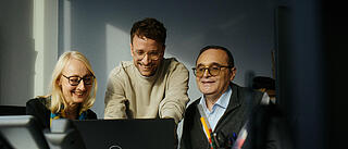 Innenaufnahme zeigt drei GESOBAU-Kolleg*innen. Die Gruppe sitzt lachend vor einem aufgeklappten Laptop; links eine blonde Frau mit dunkler Brille, mittig stehend ein jüngerer Kollege, rechts sitzend ein älterer Kollege, ebenfalls mit Brille.