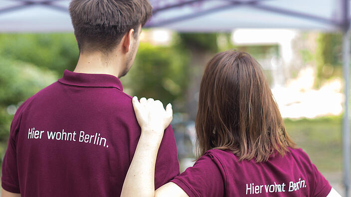 Außenaufnahme zeigt zwei Mitarbeitende der GESOBAU in den Poloshirts des Unternehmens mit dem Rücken zum Betrachter. Man liest deutlich den GESOBAU-CLaim "Hier wohnt Berlin". Die rechts stehende Mitarbeiterin hat ihren Arm auf die Schulter ihres etwas größeren Kollegen gelegt. 