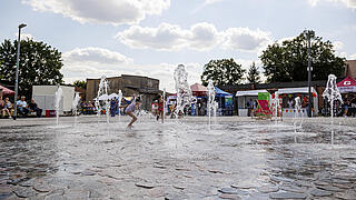 Bild zeigt das sprudelnde Wasserspiel auf dem Stadtplatz. Im Hintergrund sind spielende Kinder und weitere Besucher zu sehen.