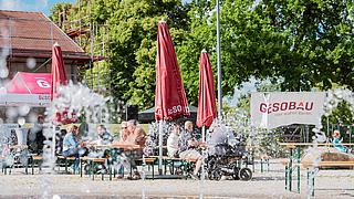 Menschen taumeln sich auf dem Stadtplatz, im Vordergrund sprudelt ein Wasserspiel