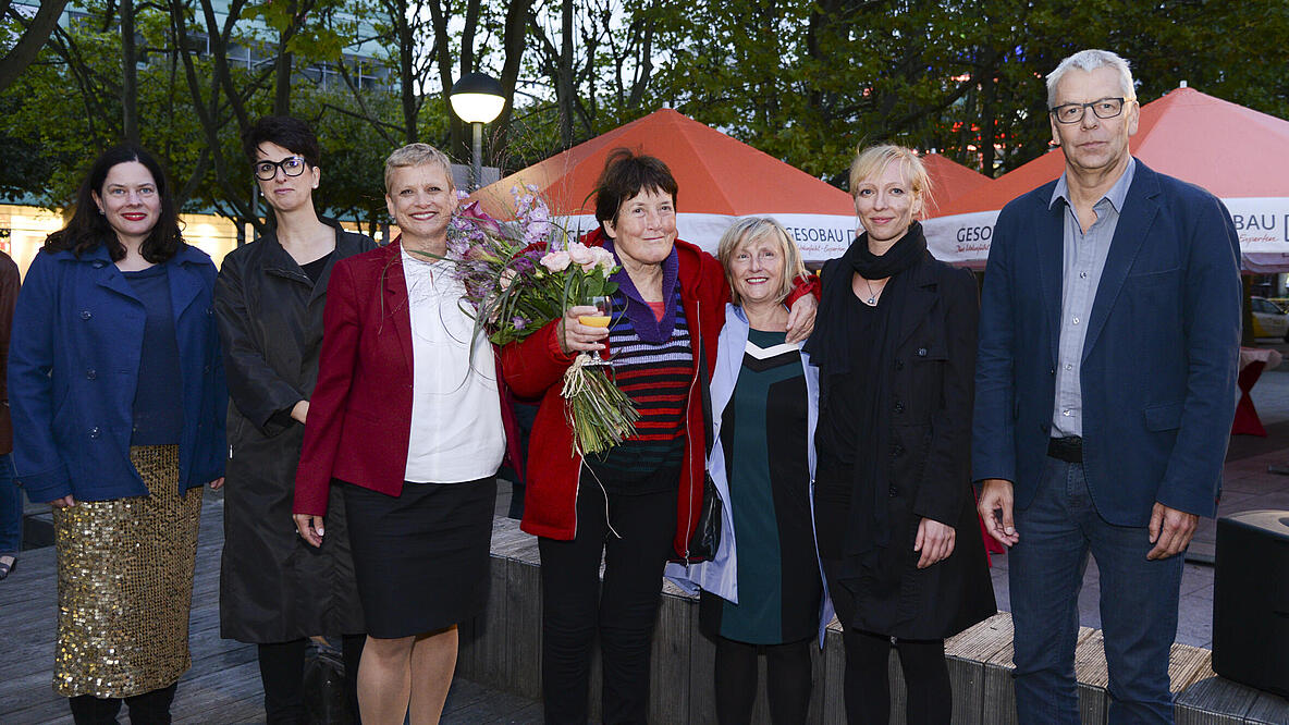 Sieben Personen nebeneinander, die Frau in der Mitte hält einen Blumenstrauß
