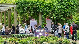 Menschen versammeln sich unter einer Pergola, die im Grünen steht.