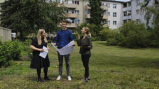 Außenaufnahme zeigt drei Mitarbeiter*innen der GESOBAU auf einer Wiese im Gespräch. Im Hintergrund der Schwyzer Kiez.