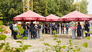Bild zeigt Menschengruppe bei Grundsteinlegung unter Sonnenschirmen. Im Hintergrund Bäume und Kräne.