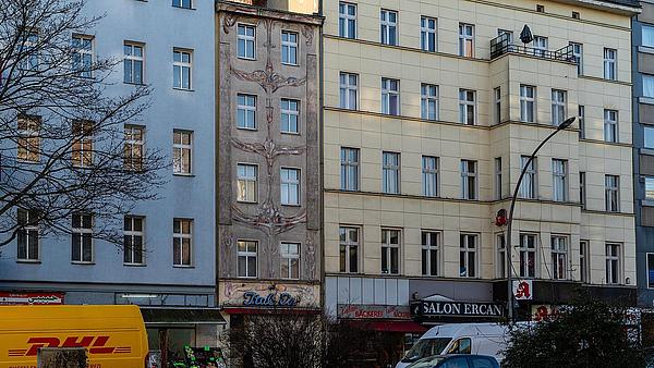 Die Außenaufnahme zeigt die Fassade einer Häuserreihe in Berlin-Wedding in der Müllerstraße. Eine blaue, graue und gelbe Fassade reihen sich aneinander. Im Erdgeschoss der Häuser befinden sich ein Bäcker, ein Restaurant und eine Apotheke. Vor den Häusern parken Autos. Auf der linken Seite lässt sich ein DHL-Lieferfahrzeug erkennen.