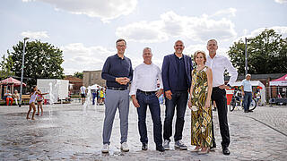 Lars Holborn und Christian Wilkens zusammen mit Staatssekretär Stephan Machulik, Bezirksstadträtin Heike Wessoly und Frank Bruckmann, Finanzvorstand der Berliner Wasserbetriebe, stehen vor dem Wasserspiel auf dem Stadtplatz. Im Hintergrund sind spielende Kinder und Besucher zu sehen.