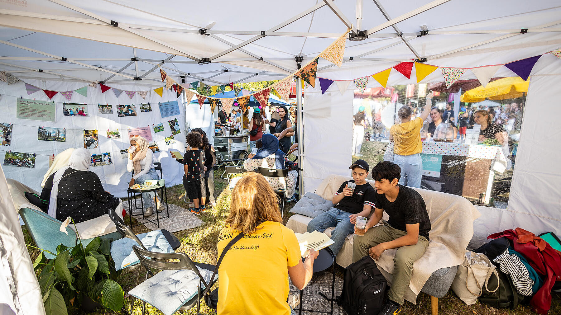 Außenaufnahme zeigt verbundende Pavillons mit bunten Wimpelgirlanden geschmückt, darin sitzend rechts im Vordergrund zwei Jugendliche im Gespräche, im Hintergrund weitere Personen.  