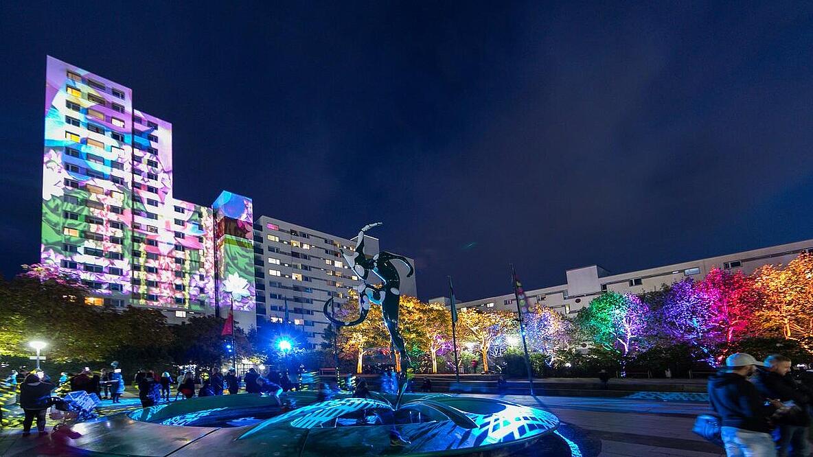 Stadtplatz mit Brunnen, rundherum Bäume und Hochhäuser, vereinzelt laufen Menschen über den Platz und alles ist bunt beleuchtet