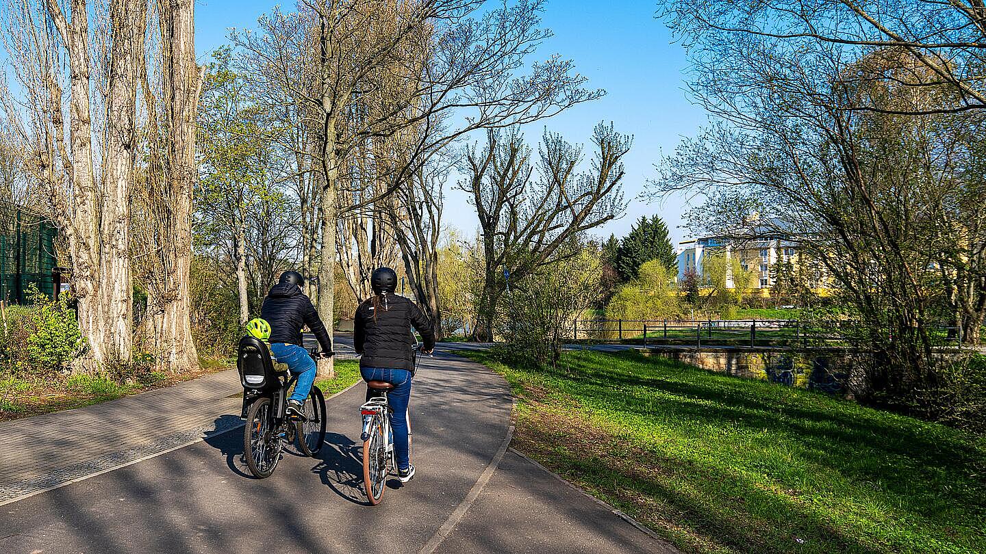 Zwei Menschen fahren einen asphaltierten Weg entlang. Auf dem Fahrrad der linken Person sitzt außerdem ein Kind im Kindersitz. Der Weg ist umgeben von grünem Rasen und Bäumen. Die Sonne scheint und es sind wenige Wolken am Himmel. Im Hintergrund rechts sieht man ein Haus in der Ferne.