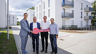Lars Holborn, Christian Wilkens, Mark Seibert und Aziz Bozkurt stehen vor den Wohnhäusern für Geflüchtete und halten gemeinsam einen großen symbolischen Schlüssel in den Händen.