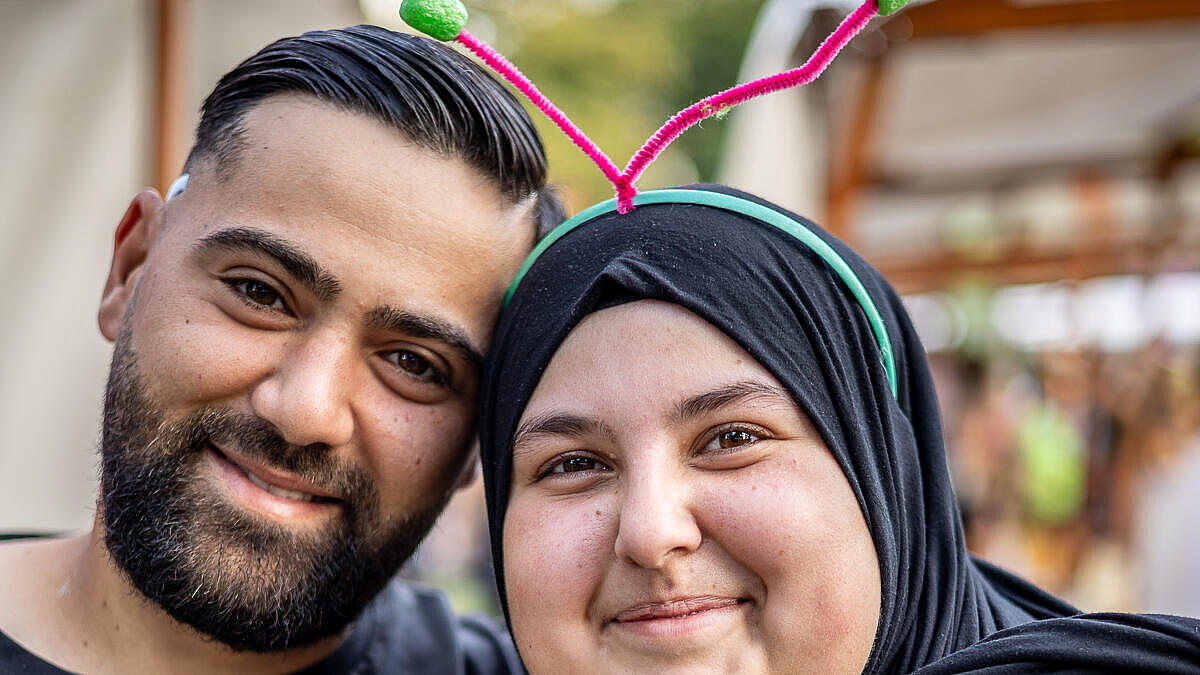 Außenaufnahme zeigt ein lächelnde Paar, recht stehend eine verschleierte Frau mit selbstgemachtem Kopfschmuck.