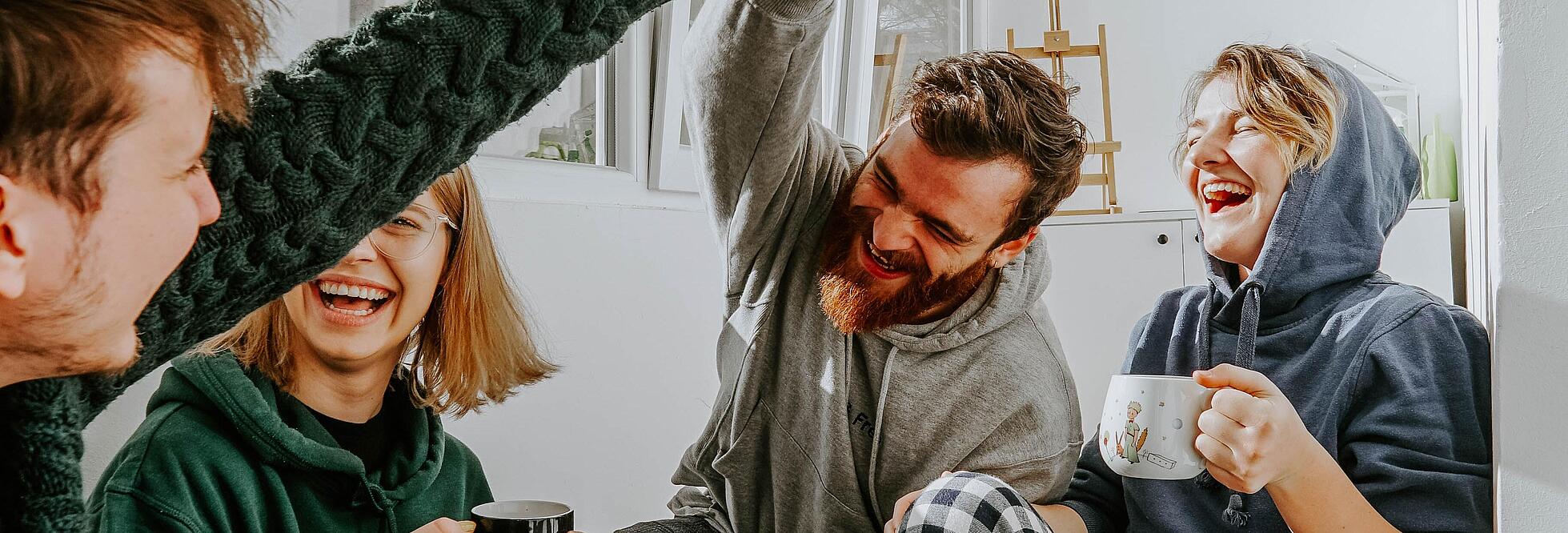 Innenaufnahme zeigt eine sitzende Gruppe von vier lachenden Menschen, die beiden Männer der Runde geben sich ein High Five.