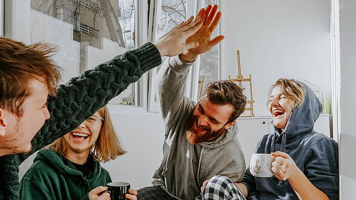 Innenaufnahme zeigt eine sitzende Gruppe von vier lachenden Menschen, die beiden Männer der Runde geben sich ein High Five.