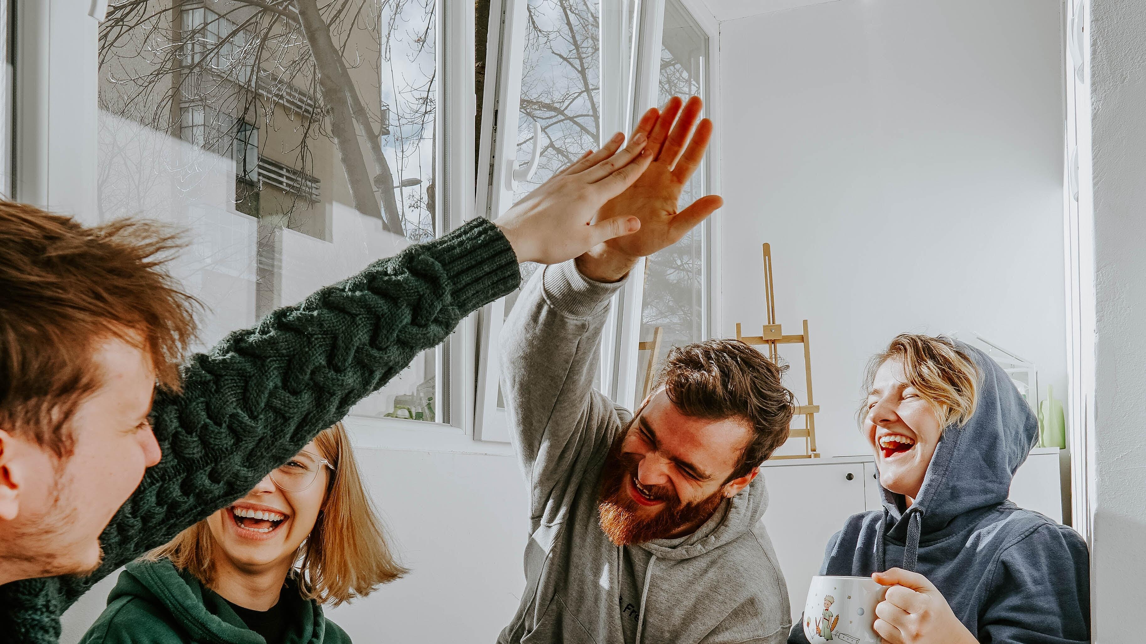 Innenaufnahme zeigt eine sitzende Gruppe von vier lachenden Menschen, die beiden Männer der Runde geben sich ein High Five.