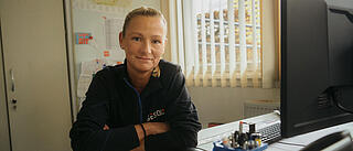 Innenaufnahme zeigt eine Hausmeisterin der GESOBAU in einem Stützpunkt an einem Arbeitsplatz sitzend. Sie hat die Arme vor sich verschränkt und schaut lächelnd in die Kamera.