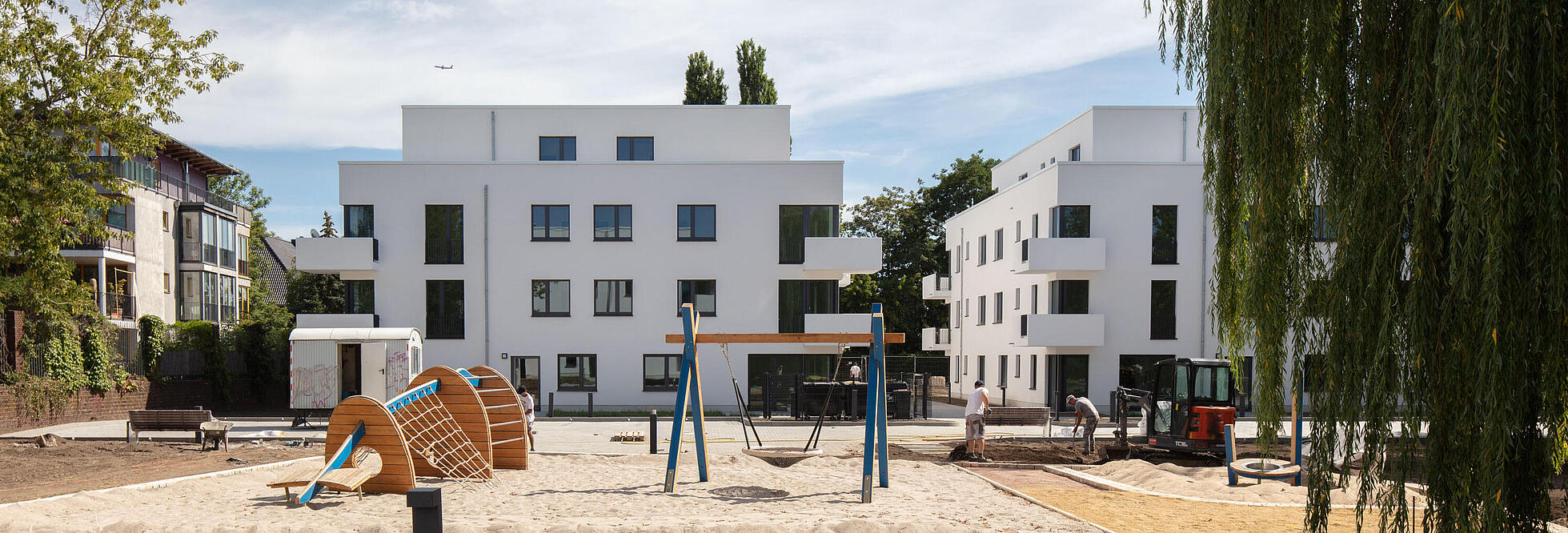 Weitwinkelaufnahme außen: Im Vordergrund ein Spielplatz mit Wipptieren, im Hintergrund in der Bildmitte zwei neu gebaute Häuser in der Abajstraße / Kastanienallee.