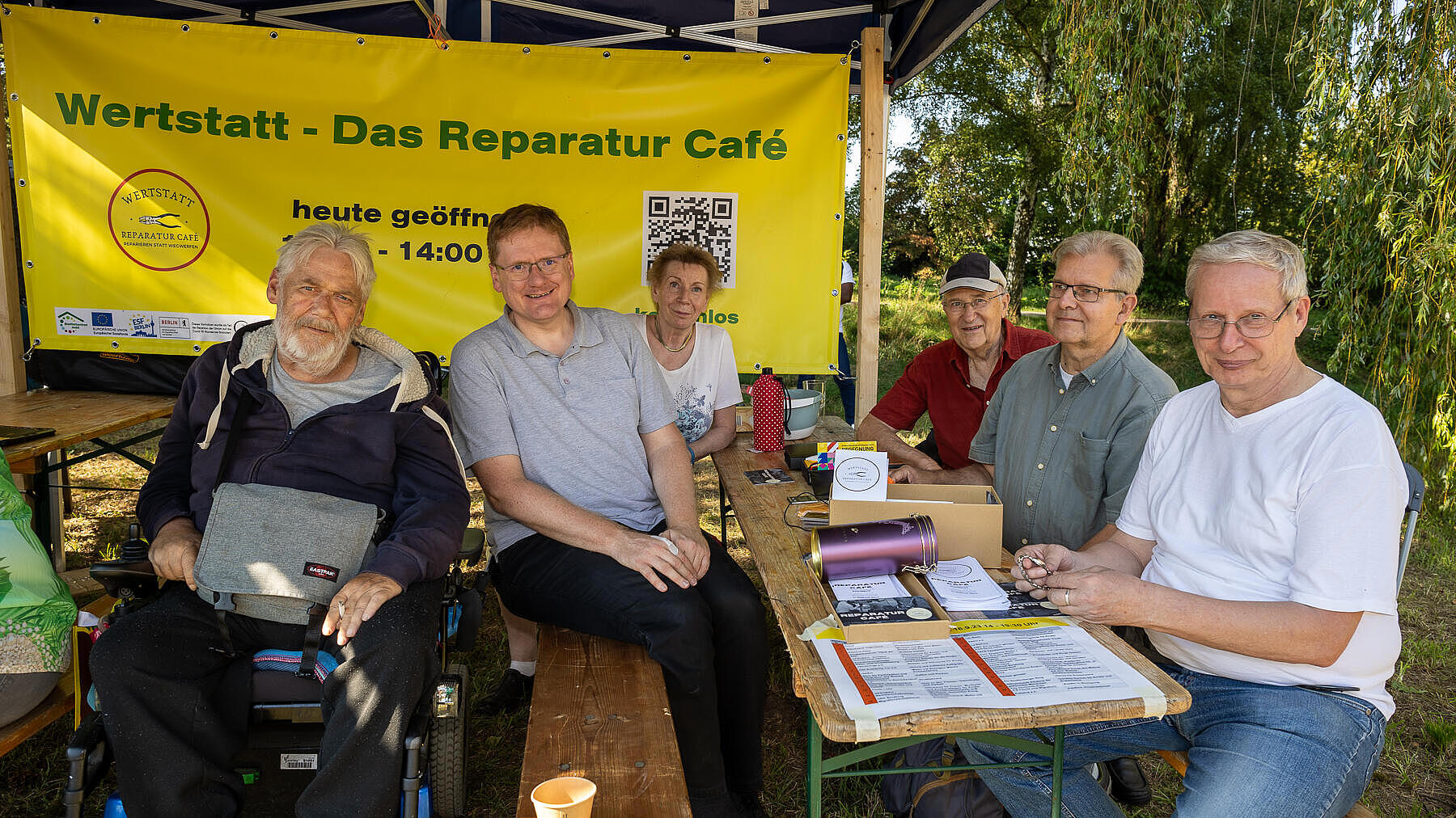 Außenaufnahme zeigt eine Gruppe von älteren Menschen, die vor einem Plakat der Wertstatt - Das Reparatur-Café sitzen.