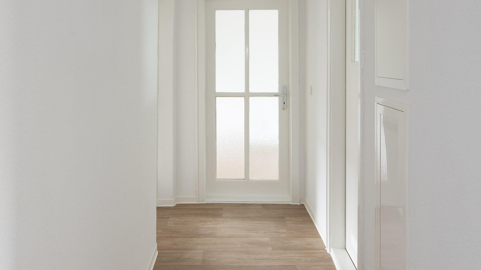 Das Bild zeigt eine modernisierte Wohnung: Ein Flur mit Holzboden, mit einer Glastür am Ende, weiße Wände, einer rechts abgehenden, weißen Zimmertür und weißen Klappen zum Stromkasten
