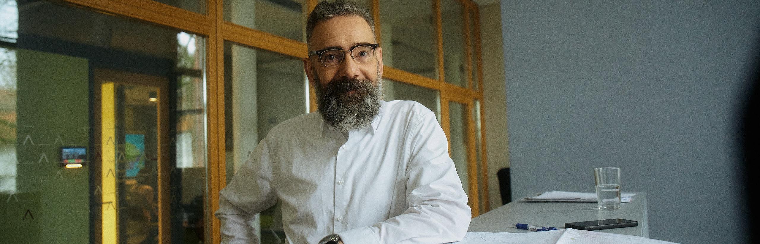 Innenaufnahme zeigt einen bebrillten Mann um die 50 mit kurzen Haaren und gepflegtem Vollbart, der, mit dem linken Ellbogen auf ein Regal und dem rechten Arm in seine Hüfte gestützt, lächelnd in die Kamera blickt. Vor ihm liegen unscharf erkennbar ein Mobiltelefon, ein Büroschlüssel und ein aufgefalteter Plan.