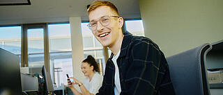 Innenaufnahme zeigt zwei Mitarbeitende der GESOBAU bei der Arbeit. Der Kollege im Vordergrund sitz an einem Schreibtisch und lächelt in die Kamera.