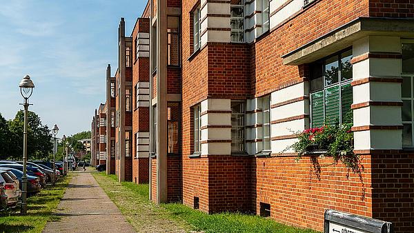 Die Außenaufnahme zeigt auf der rechten Seite die Fassade eines Wohnhauses im Schillerpark, Berlin-Wedding. Das Gebäude besteht aus rotem Backstein und die Fenster sind teilweise mit Blumen beschmückt. Ein Gehweg führt entlang des Hauses, links davon parkende Autos und Laternen.
