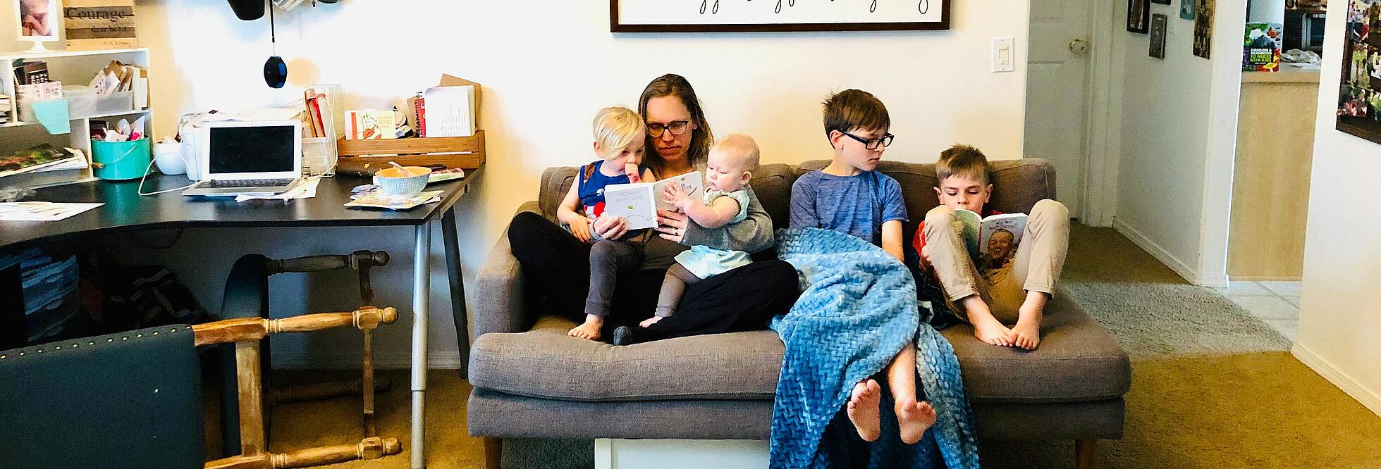 Innenaufnahme zeigt eine Familie bestehend aus einer Frau und vier Kindern unterschiedlichen Alters in einem offenen Wohnzimmer. Sie sitzend gemeinsam auf einem Sofa, die Frau hat je ein Kleinkind auf einem Bein im Schoß sitzend und schaut mit ihnen ein Buch an. Die beiden älteren Kinder lesen ebenfalls ein Buch.
