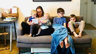 Innenaufnahme zeigt eine Familie bestehend aus einer Frau und vier Kindern unterschiedlichen Alters in einem offenen Wohnzimmer. Sie sitzend gemeinsam auf einem Sofa, die Frau hat je ein Kleinkind auf einem Bein im Schoß sitzend und schaut mit ihnen ein Buch an. Die beiden älteren Kinder lesen ebenfalls ein Buch.
