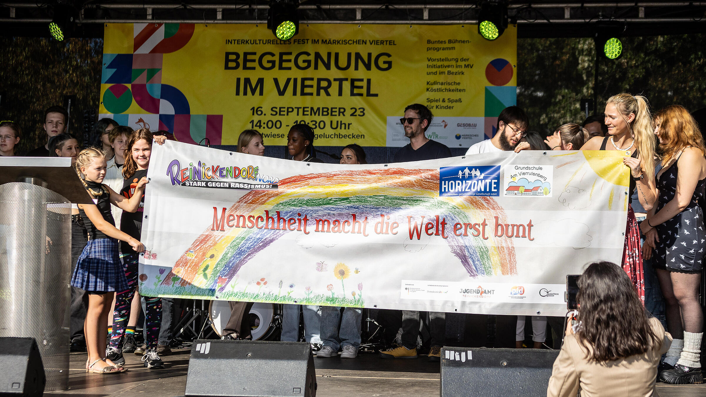 Außenaufnahme zeigt eine Gruppe vpn Jugendlichen, die auf einer Bühne ein Banner mit der Aufschrift "Menschheit macht die Welt erst bunt" hochalten.