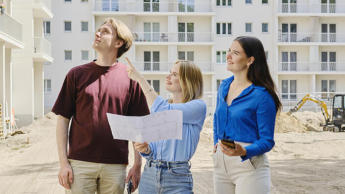 Außenaufnahme zeigt drei Auszubildende auf einer Baustelle, die mittig stehende Person hält einen Plan in der Hand, alle drei schauen nach oben, offenkundig im Begriff, etwas mit dem Plan abgleichend.