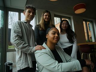 Innenaufnahme zeigt junge Auszubildende, die sitzend selbstbwusst in die Kamera lächelt. Hinter ihr drei weitere, gleichalttrige Personen, zwei junge Frauen mittig und rechts, linker Hand ein Kollege, der seine Hand auf die Schulter der sitzenden KOllegin gelegt hat. Alle vier lächeln leicht und selbstbewusst.