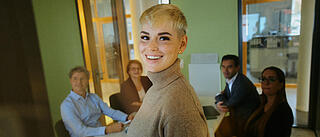 Innenaufnahme zeigt eine junge Frau mit Kurzhaarschnitt, die lachend und sympathisch in die Kamera blickt. Im Hintergrund sitzend vier weitere Kolleg*innen, die auf das Smartboard neben der ersten Person schauen.