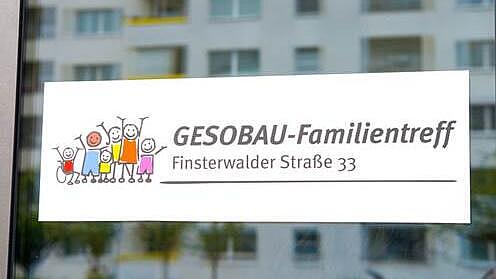 Blick auf eine Scheibe des GESOBAU-Familientreffs mit de Schild in der Mitte, dahinter spiegeln sich die Hausfassaden.