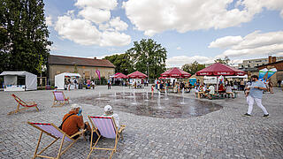 Bild zeigt das sprudelnde Wasserspiel auf dem Stadtplatz. Im Hintergrund sind spielende Kinder und weitere Besucher zu sehen.