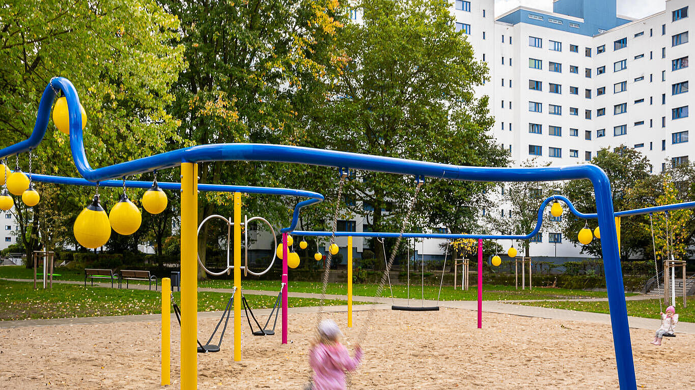 Bild zeigt neue Spielfläche im Märkischen Viertel, im Vordergrund Kind auf Schaukel, im Hintergrund Bäume und Wohnhäuser.