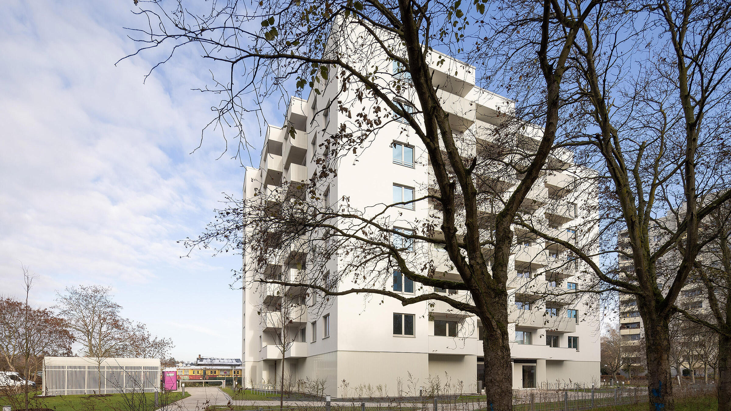 Seitlicher Blick auf den Neubau in der Finsterwalder Straße, im Vordergrund Bäume. Linkseitig der eingezäunte Müllplatz.