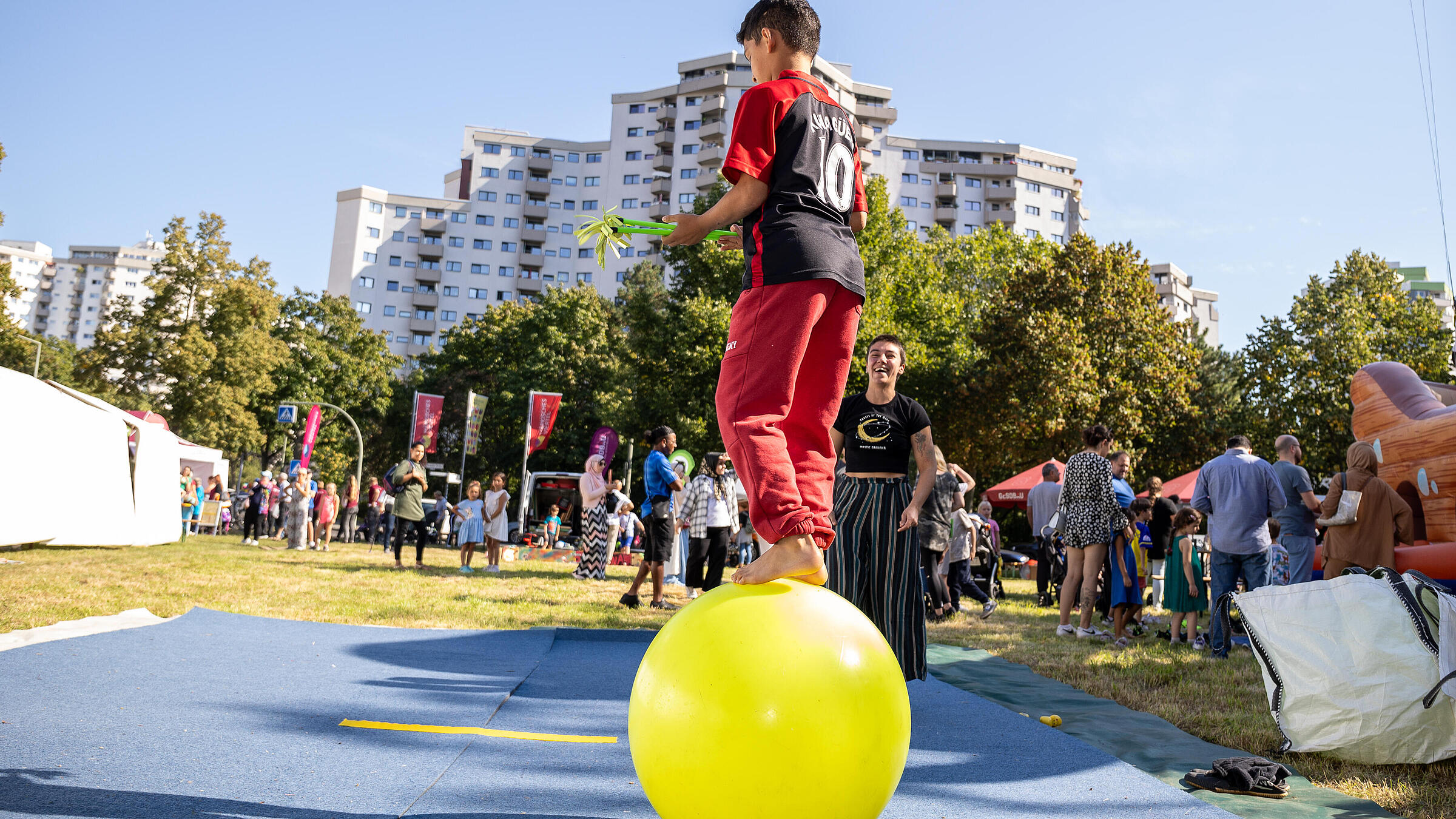 Außenaufnahme zeigt ein Kind mit dem Rücken zum Betrachter, das auf einem gelben Turnball balanciert. Im Hintergrund die Kulisse der markanten Hochhäuser vom Märkischen Viertel.
