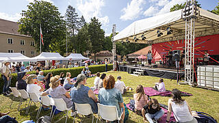 Weitwinkelaufnahme zeigt Zuschauer*innen vor der Kleinen Bühne im Sonnenschein.