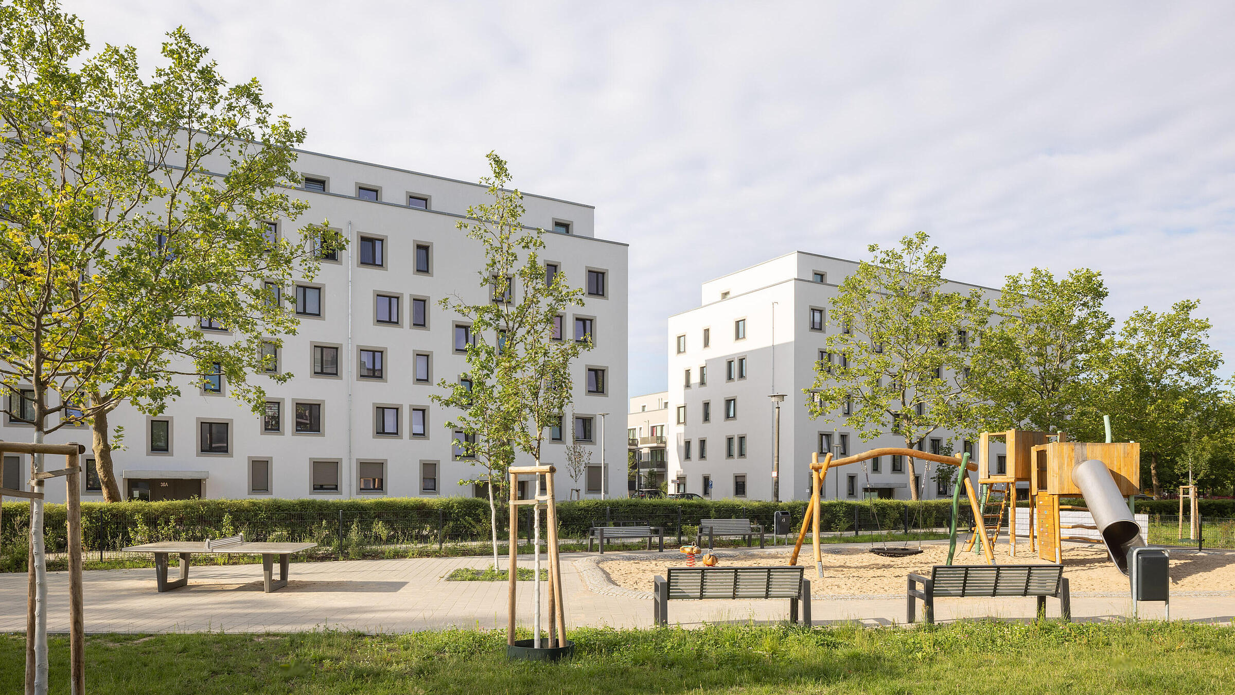 Stadtpark im Quartier Stadtgut Hellersdorf: Grünfläche umgeben von Bänken und einer Tischtennisplatze; im Hintergrund sind ein Spielplatz und zwei moderne Wohnhäuser zu sehen.