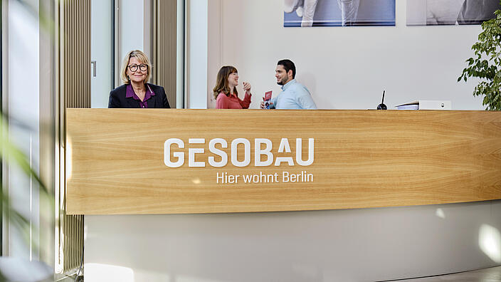 Innenaufnahme zeigt eine Personengruppe, zwei wieblich und eine männlich gelesene Person, hinter dem Tresen der GESOBAU stehend. DIe weibliche Person ganz links lächelt in die Kamera, die beiden anderen Personen sind im Gespräch miteinander.