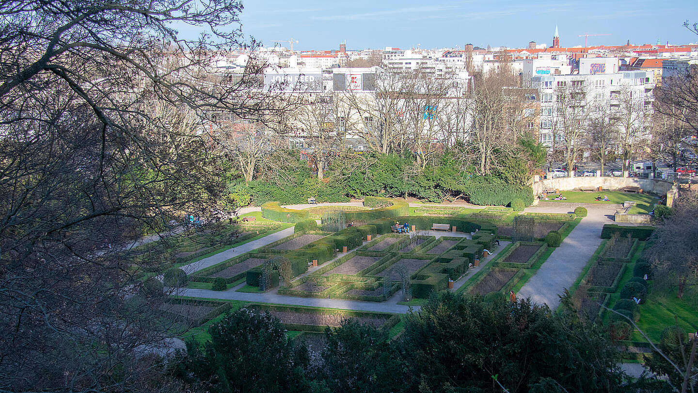 Die Außenaufnahme zeigt den Volkspark Humboldthain in Berlin-Gesundbrunnen mit grünen Wiesen, Bäumen und Spazierwegen. Im Hintergrund sind man ein Dächermeer Berlins. 