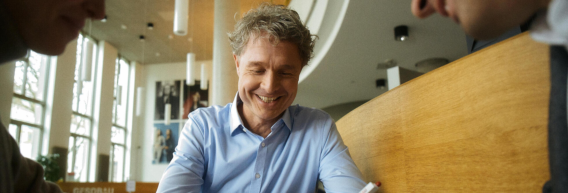 Innenaufnahme zeigt drei um einen Tisch sitzende Personen. Die rechts sitzende, männliche Person, hält einen Stift in der Hand und liest ein vor ihr aufgeschlagenes Dokument. Mittig sitz einelachende, männliche Person, die offenbar beim Signieren hilft und das Paar berät. 