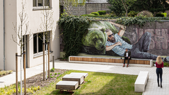 Außenaufnahme zeigt den begrünten HInterhof-Standort des GESOWORX in der Langhansstraße 22. Die Grundstücksmauer ist mit dem Mural eines halb liegenden Hipsters und einem grünen Berliner Bär besprüht.