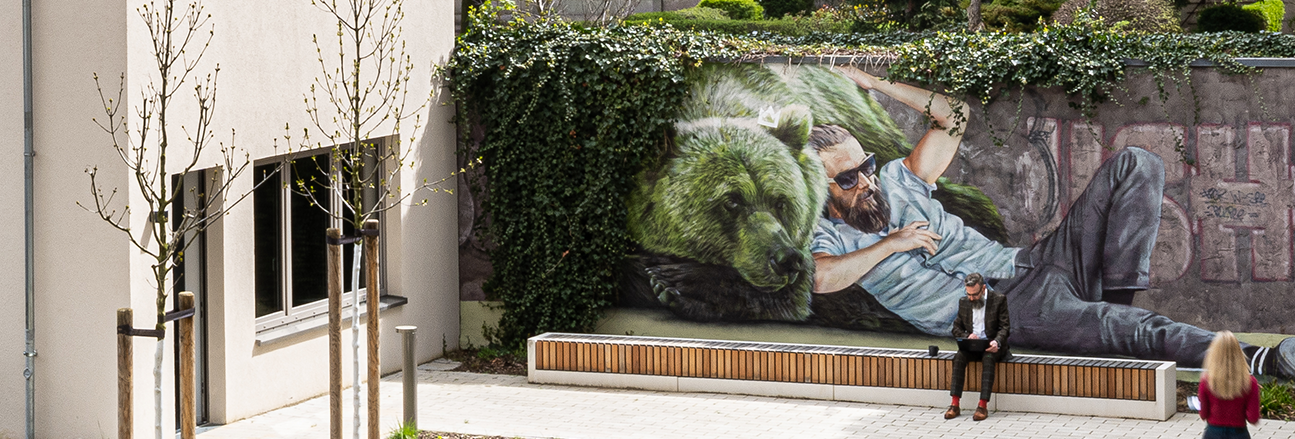 Außenaufnahme zeigt den begrünten HInterhof-Standort des GESOWORX in der Langhansstraße 22. Die Grundstücksmauer ist mit dem Mural eines halb liegenden Hipsters und einem grünen Berliner Bär besprüht.