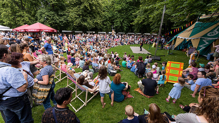 Außenaufnahme zeigt die Wiese am Schloss Niederschönhausen beim 21. Kunstfest in Pankow. Sie ist mit vielen Zuschauer*innen, die halbkreisförmig um ein Zirkuszelt auf dem Boden sitzen und sich eine Vorstellung ansehen.