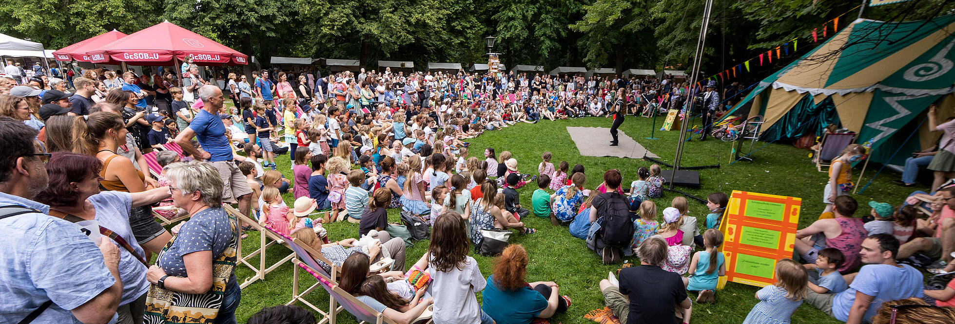 Außenaufnahme zeigt die Wiese am Schloss Niederschönhausen beim 21. Kunstfest in Pankow. Sie ist mit vielen Zuschauer*innen, die halbkreisförmig um ein Zirkuszelt auf dem Boden sitzen und sich eine Vorstellung ansehen.