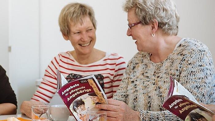 Zwei ältere Damen sitzen an einem Tisch, eine hält ein Buch und sie unterhalten sich lächelnd.