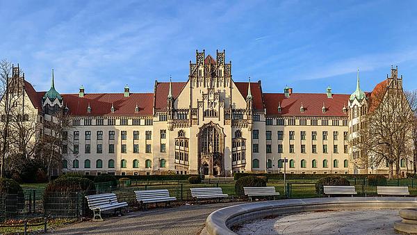 Die Außenaufnahme zeigt den Brunnenplatz in Berlin Wedding. Im rechten Vordergrund befindet sich der Ausschnitt eines großen, runden Brunnens, der nicht mit Wasser gefüllt ist. Um den Brunnen herum befinden sich, ebenfalls im Kries aufgestellt, mehrere Parkbänke. Im Hintergrund befindet sich eine umzäunte Wiese und dahinter ein großes, prunkvolles Gebäude.