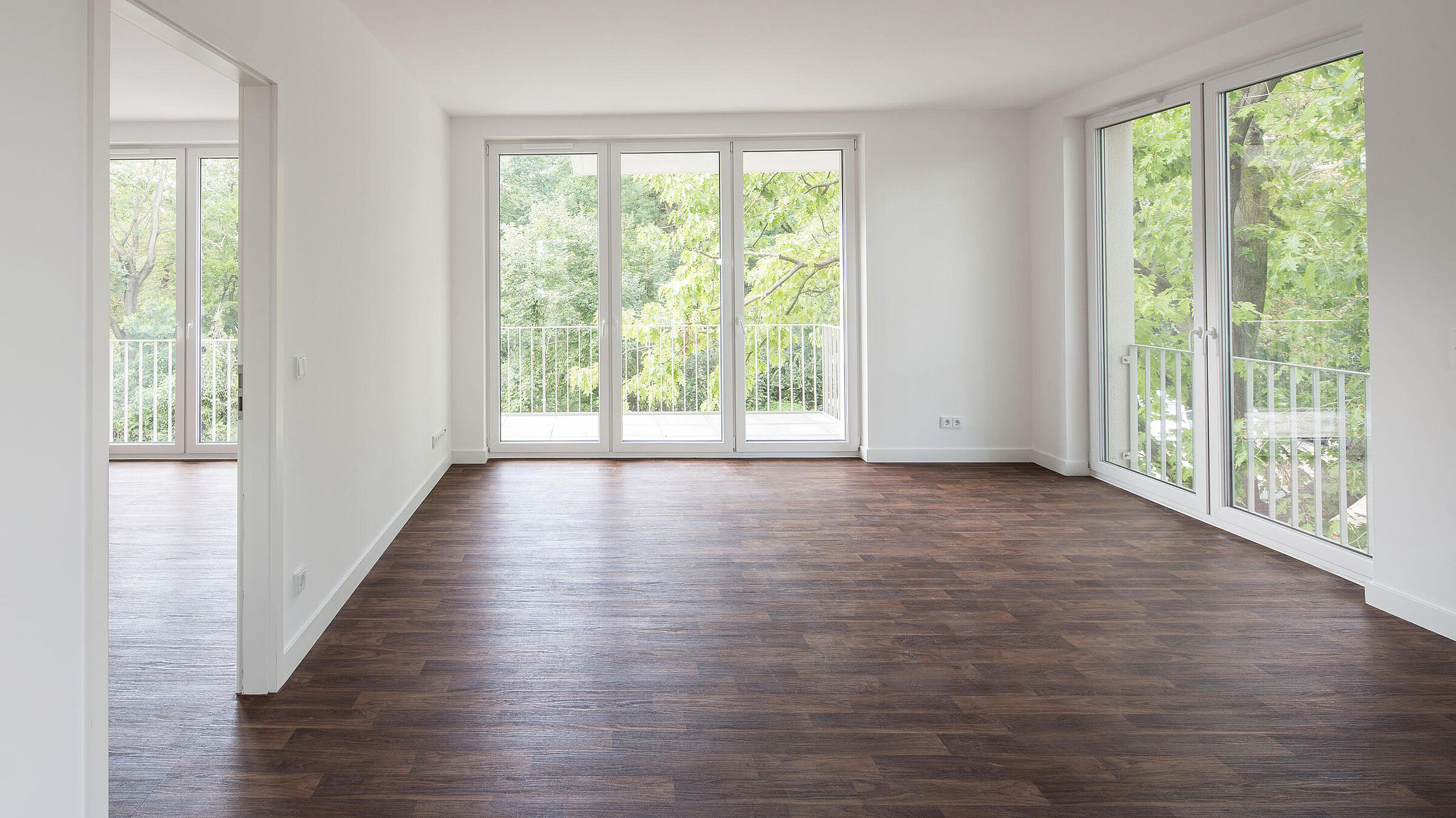 Innenaufnahme zeigt ein leeres Zimmer mit bodentiefen Fenstern, einem Fußboden in Holzoptik und Balkon.