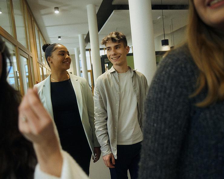 Innenaufnahme zeigt eine Gruppe von vier Auszubildenden bzw. Studierenden in Bewegung, unscharf und unvollständig im Vordergrund erkennbar, zwei lachende junge Frauen. Mittig und deutchlich sichtbar links eine junge Frau und rechts ein junger Mann, lächelnd im Gespräch.
