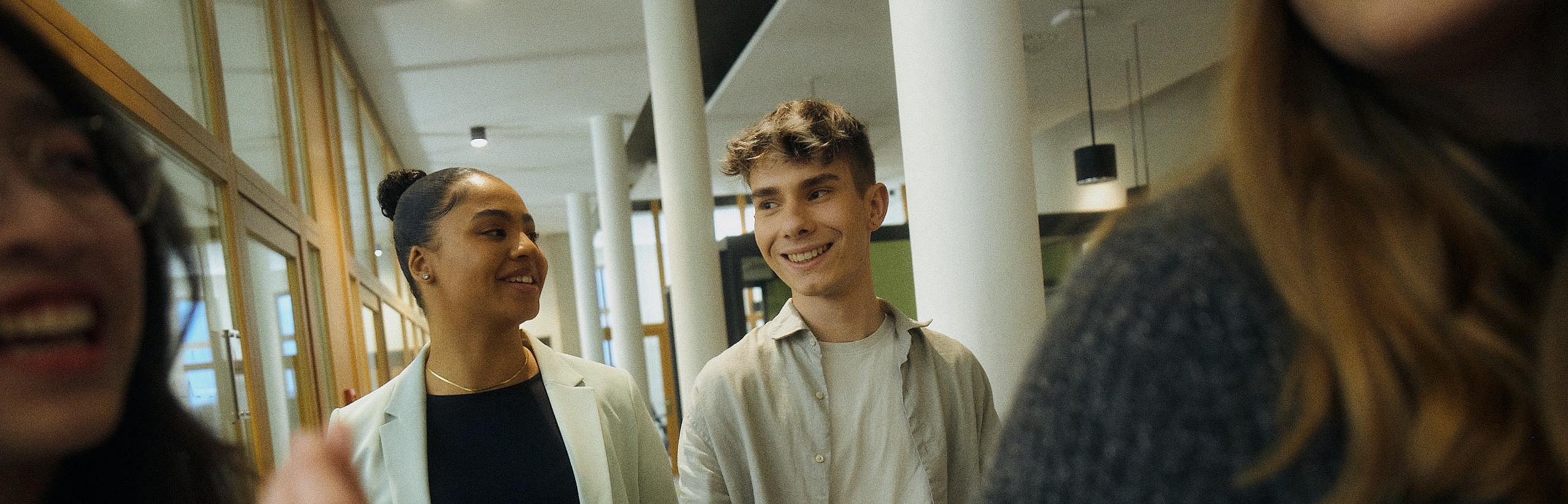 Innenaufnahme zeigt eine Gruppe von vier Auszubildenden bzw. Studierenden in Bewegung, unscharf und unvollständig im Vordergrund erkennbar, zwei lachende junge Frauen. Mittig und deutchlich sichtbar links eine junge Frau und rechts ein junger Mann, lächelnd im Gespräch.